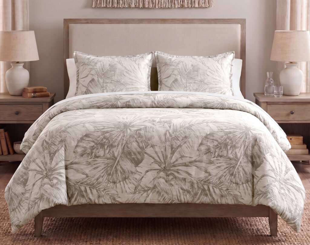 Bed with a beige upholstered headboard, palm-pattern duvet, and two matching pillows on wooden nightstands with lamps on each side.