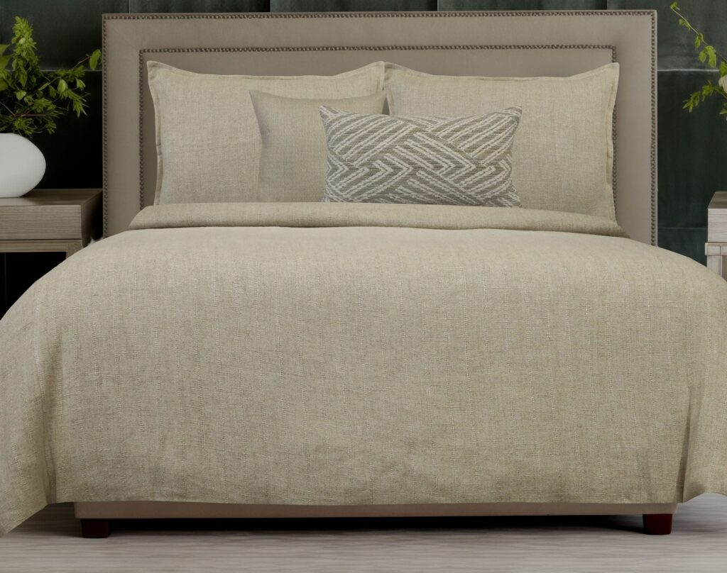 Beige bed with a cushioned headboard, three beige pillows, and a patterned gray lumbar pillow on a matching duvet.