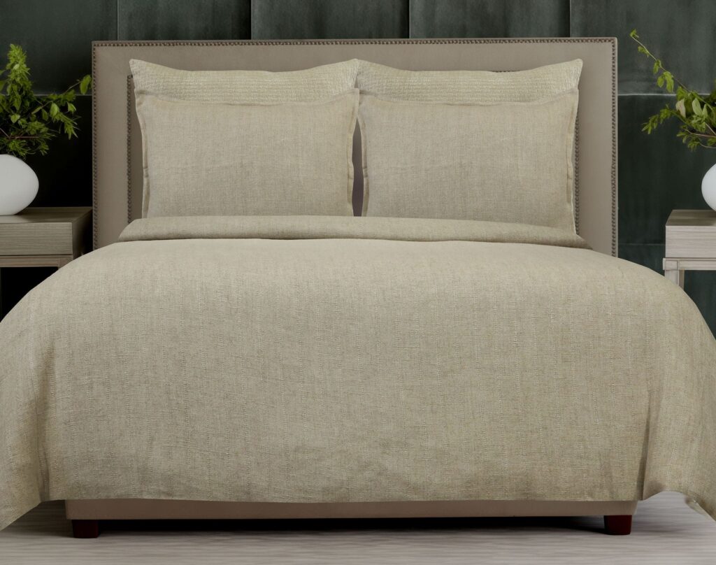 Beige upholstered bed with two matching pillows and textured bedding in a neutral, modern bedroom. A nightstand and greenery flank the bed.