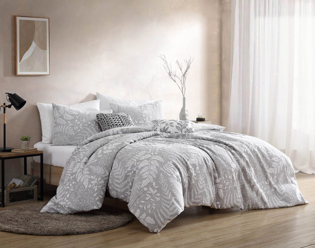 Bed with grey botanical-patterned bedding and matching pillows, beside a small side table with a lamp and plant in a softly lit bedroom.