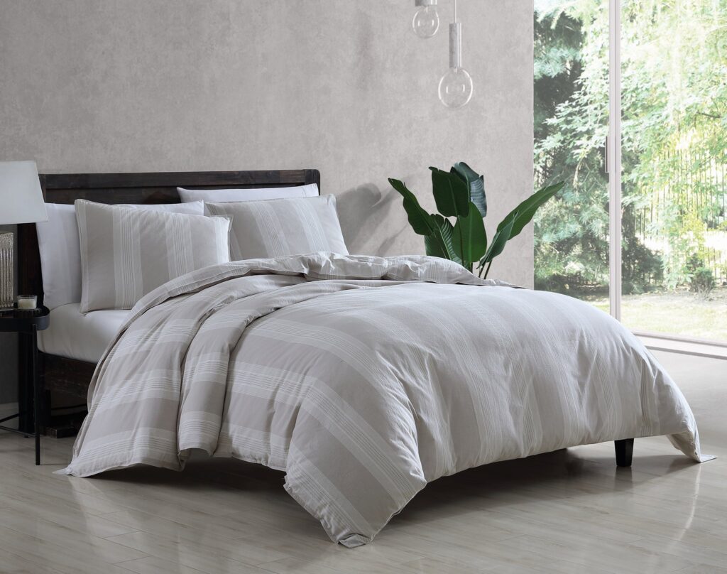 Neatly made bed with gray striped bedding and white pillows, a dark wood headboard, and a green potted plant by a large window.