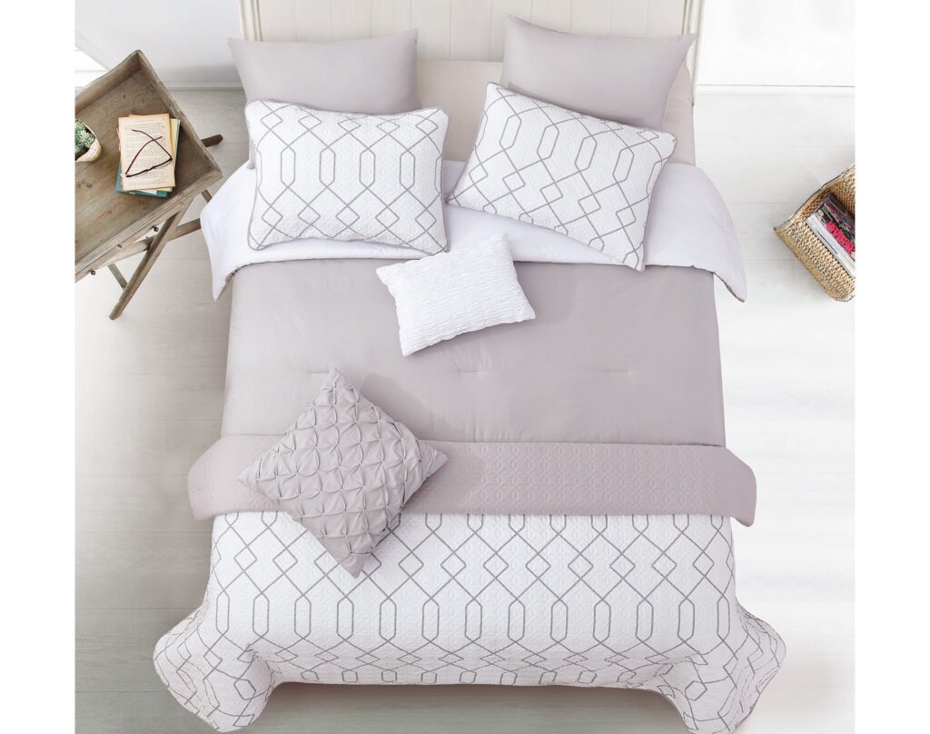 Top-down view of a pale gray bed with a geometric quilt, matching pillows, and a textured throw; a wooden side table with books on the left and a wicker magazine basket on the right.