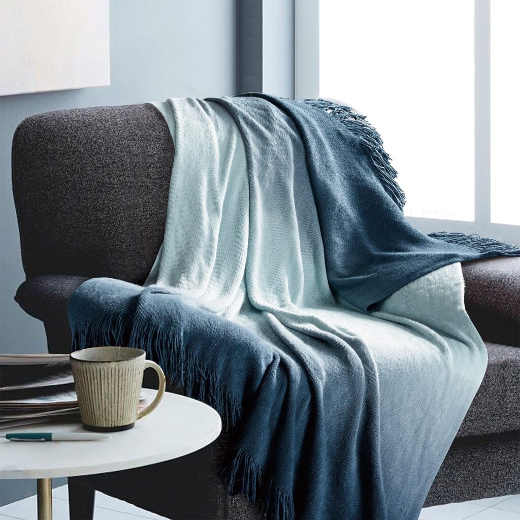 Cozy living room scene with a grey sofa draped in light and dark blue blankets and a mug on a white round table nearby.