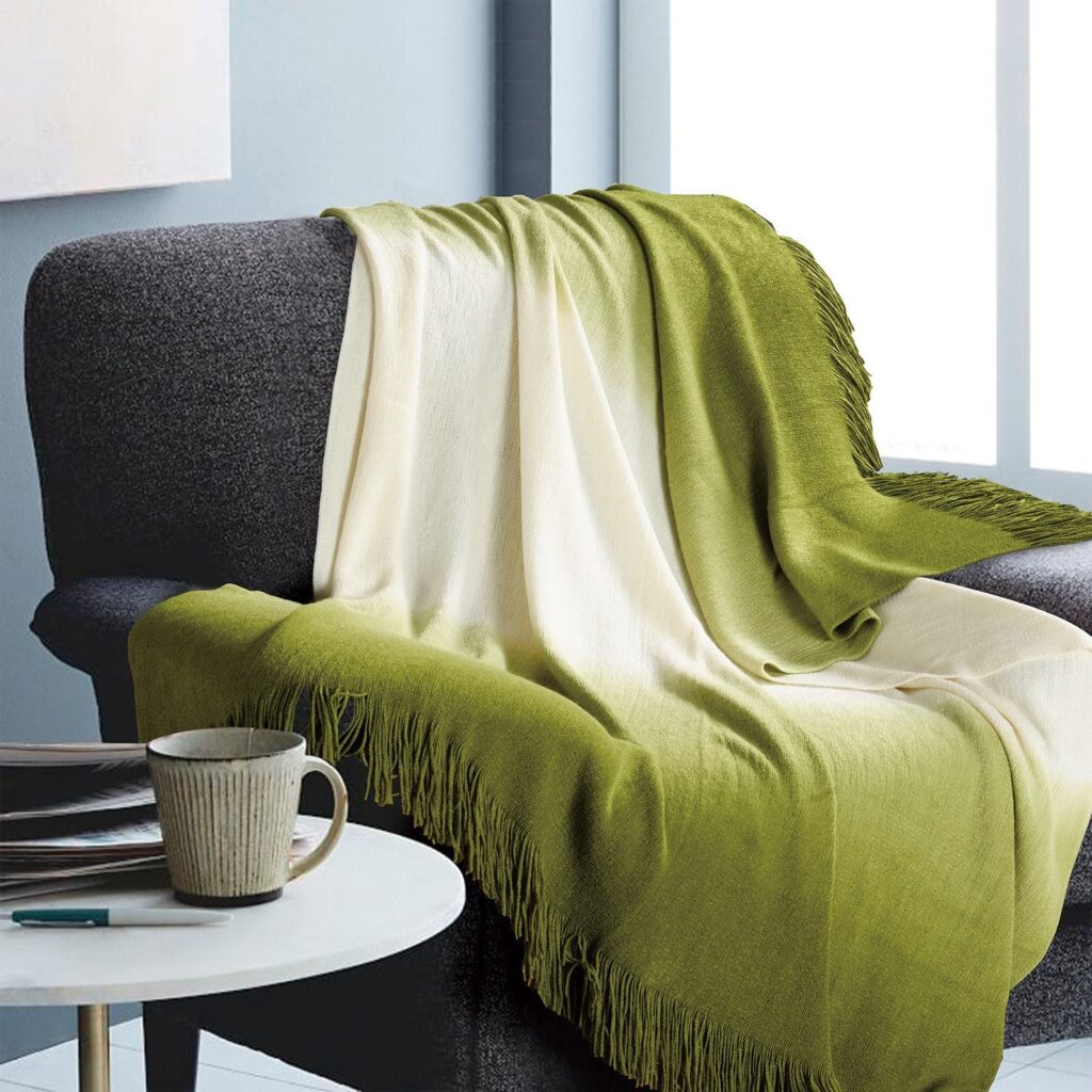 Grey sofa draped with olive green and cream throws, fringe along the edges, with a mug on a white round table nearby in a bright living room.