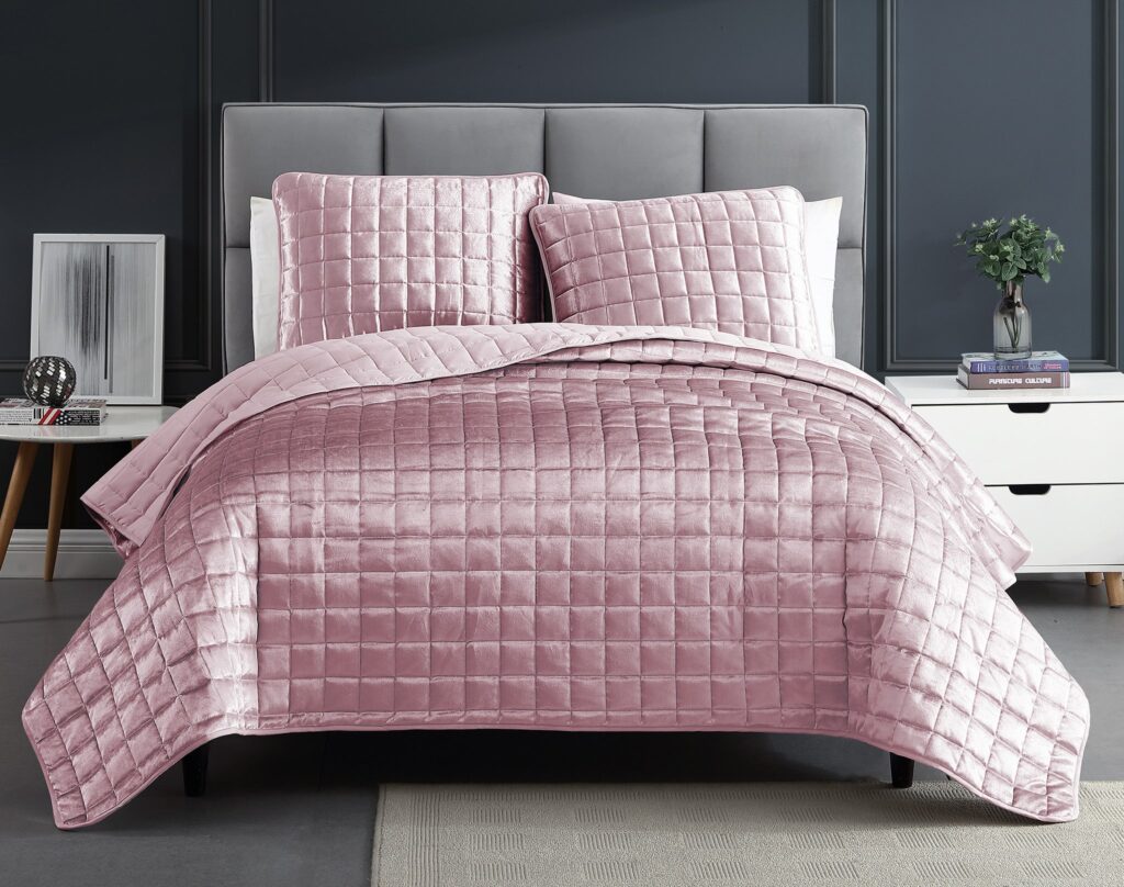 Bed with pink quilted comforter and matching pillows against a grey upholstered headboard in a dark-toned bedroom.