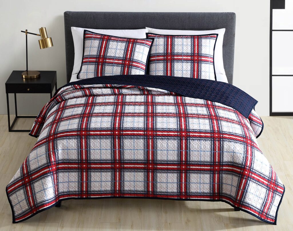 Bedroom scene featuring a bed with a red, white, and navy plaid quilt, white pillows, and a black nightstand with a brass lamp beside it.