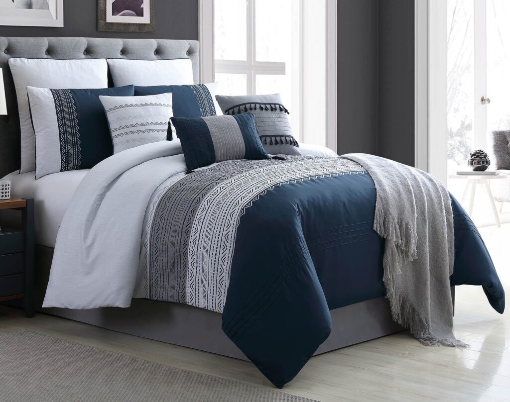 Queen bed with navy and white patterned duvet and tufted gray headboard, multiple decorative pillows, and soft fringe throw in a modern bedroom.