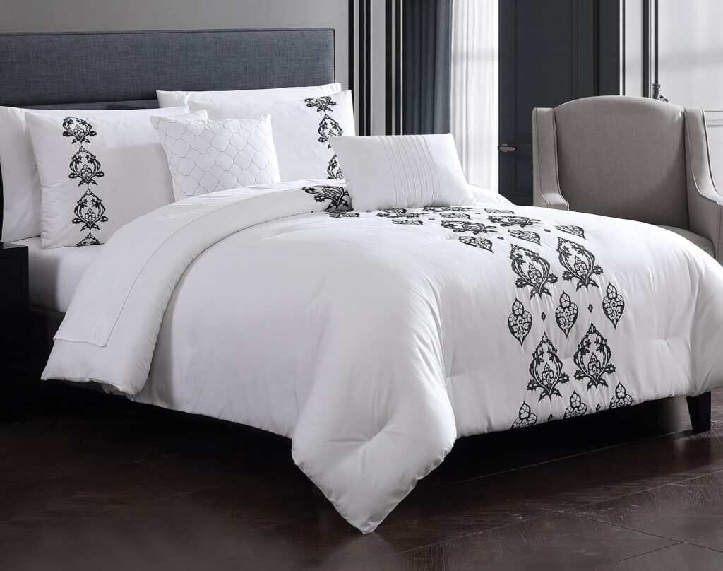 Neatly made bed in a modern bedroom with white linens, black embroidered patterns, and a blue upholstered headboard.